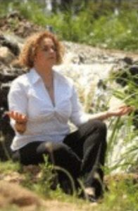 יעל יושבת ליד מעיין עם כפות ידיה מופנות כלפי מעלה בתפילה או במדיטציה